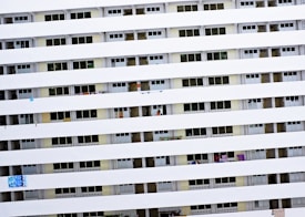 A tall residential building with multiple rows of uniform balconies. Each balcony is enclosed with rails, and some have laundry or small items hanging. The facade is primarily white with consistent horizontal lines formed by the balcony railings.