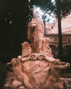 A classical stone sculpture featuring several figures, including a central robed figure standing on a pedestal surrounded by cherubs. Two muscular male figures kneeling on the sides appear to be holding the pedestal. A bird is perched on the head of the central figure. The background involves lush greenery and a historic building with arches.