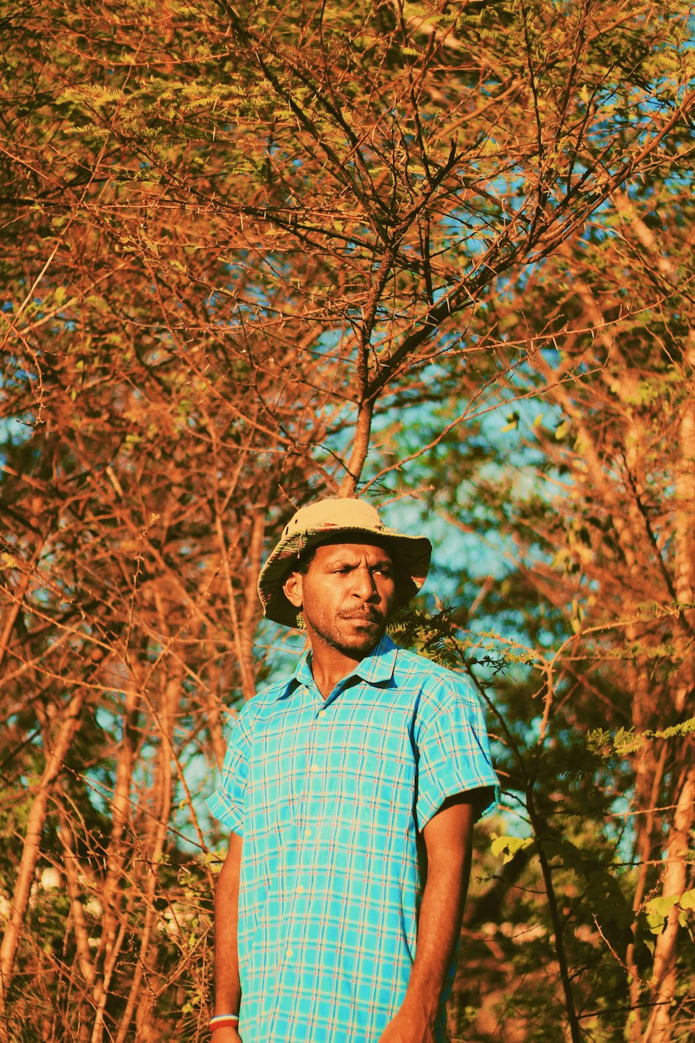 Hombre con camisa abotonada a cuadros azules y blancos con sombrero marrón de pie cerca de la hoja marrón