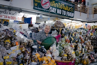 An older woman stands behind a market stall overflowing with various packaged goods. The stall is filled with a wide assortment of dried foods, fruits, nuts, and spices, all contained in plastic bags. A green plastic basket is held in front of her. Overhead, a sign in multiple languages advocates against the use of foam.
