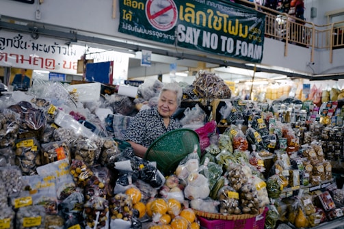 An older woman stands behind a market stall overflowing with various packaged goods. The stall is filled with a wide assortment of dried foods, fruits, nuts, and spices, all contained in plastic bags. A green plastic basket is held in front of her. Overhead, a sign in multiple languages advocates against the use of foam.