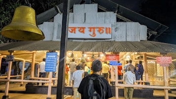 A group of people are gathered in front of a temple-like structure with a large bell hanging prominently. The building has a corrugated metal roof and a sign with Hindi script above the entrance. The scene is lit by artificial lights, suggesting it is evening or night. Several informational signs are displayed around the entrance.