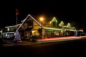 A charming, well-lit bakery with a traditional architectural style, adorned with festive lights. There are Christmas trees outside, decorated with colorful lights, and the scene includes the blur of car lights passing by, indicating movement. The area is softly illuminated by streetlights, creating a warm ambiance.