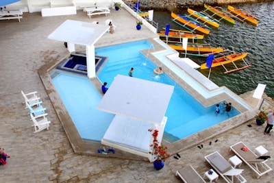 An aerial view of a modern outdoor swimming pool complex next to a body of water. The pool is divided into sections with several people enjoying the water. The area is surrounded by white lounge chairs and umbrellas, and nearby there are colorful wooden boats docked at the water's edge. The setting conveys a relaxed and recreational atmosphere, with a harmonious blend of cool blues and warm natural tones.