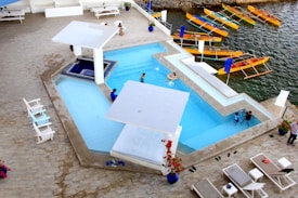 An aerial view of a modern outdoor swimming pool complex next to a body of water. The pool is divided into sections with several people enjoying the water. The area is surrounded by white lounge chairs and umbrellas, and nearby there are colorful wooden boats docked at the water's edge. The setting conveys a relaxed and recreational atmosphere, with a harmonious blend of cool blues and warm natural tones.