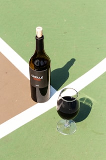 A bottle of red wine labeled 'ONDULÉ' with a cork marked '2013' is positioned on a textured surface intersected by white and brown lines. Next to it is a glass filled with red wine, casting a distinct shadow.