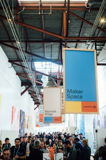 A bustling indoor corridor filled with people, many of whom appear to be engaged in conversation or moving through the space. Above them, large colorful signs direct attendees to different areas such as the Maker Space, Keynote Stage, and WC. Natural light filters from skylights, casting a bright ambiance.