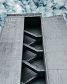 A large, concrete building with the words 'Faculty of Engineering' inscribed at the top. The building features an external staircase zigzagging up the facade, visible through a tall, narrow vertical opening. The sky is partly cloudy, providing a dramatic backdrop.