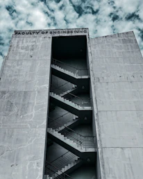 A large, concrete building with the words 'Faculty of Engineering' inscribed at the top. The building features an external staircase zigzagging up the facade, visible through a tall, narrow vertical opening. The sky is partly cloudy, providing a dramatic backdrop.
