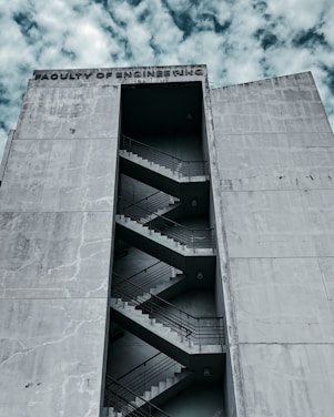 A large, concrete building with the words 'Faculty of Engineering' inscribed at the top. The building features an external staircase zigzagging up the facade, visible through a tall, narrow vertical opening. The sky is partly cloudy, providing a dramatic backdrop.