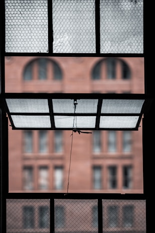 An open window with a wire mesh screen reveals a blurred view of a brick building with arched windows in the background. The window is held open by a cord attached to a lever.