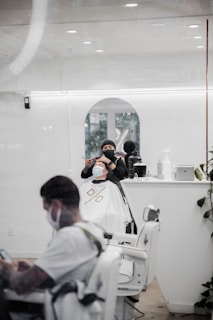 A barber wearing a mask and dark clothing cuts a customer's hair in a well-lit, modern barbershop. The customer, also wearing a mask, is seated in a white cape. Another person waits in the foreground, looking at a phone. The interior is sleek with white walls, large mirrors, and green plants.