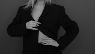 A person wearing an oversized black blazer partially conceals their torso. The image, in grayscale, highlights the minimalist and sophisticated style. The fingers of one hand are visible, gently adjusting the lapel of the blazer.