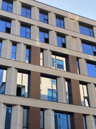 A modern office building with a grid-like pattern of windows and facade panels. The facade consists of alternating sections of beige and brown, with reflective glass windows that mirror parts of the building and the sky. The design features clean, straight lines and geometric shapes.