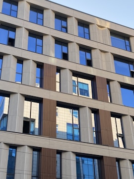 A modern office building with a grid-like pattern of windows and facade panels. The facade consists of alternating sections of beige and brown, with reflective glass windows that mirror parts of the building and the sky. The design features clean, straight lines and geometric shapes.