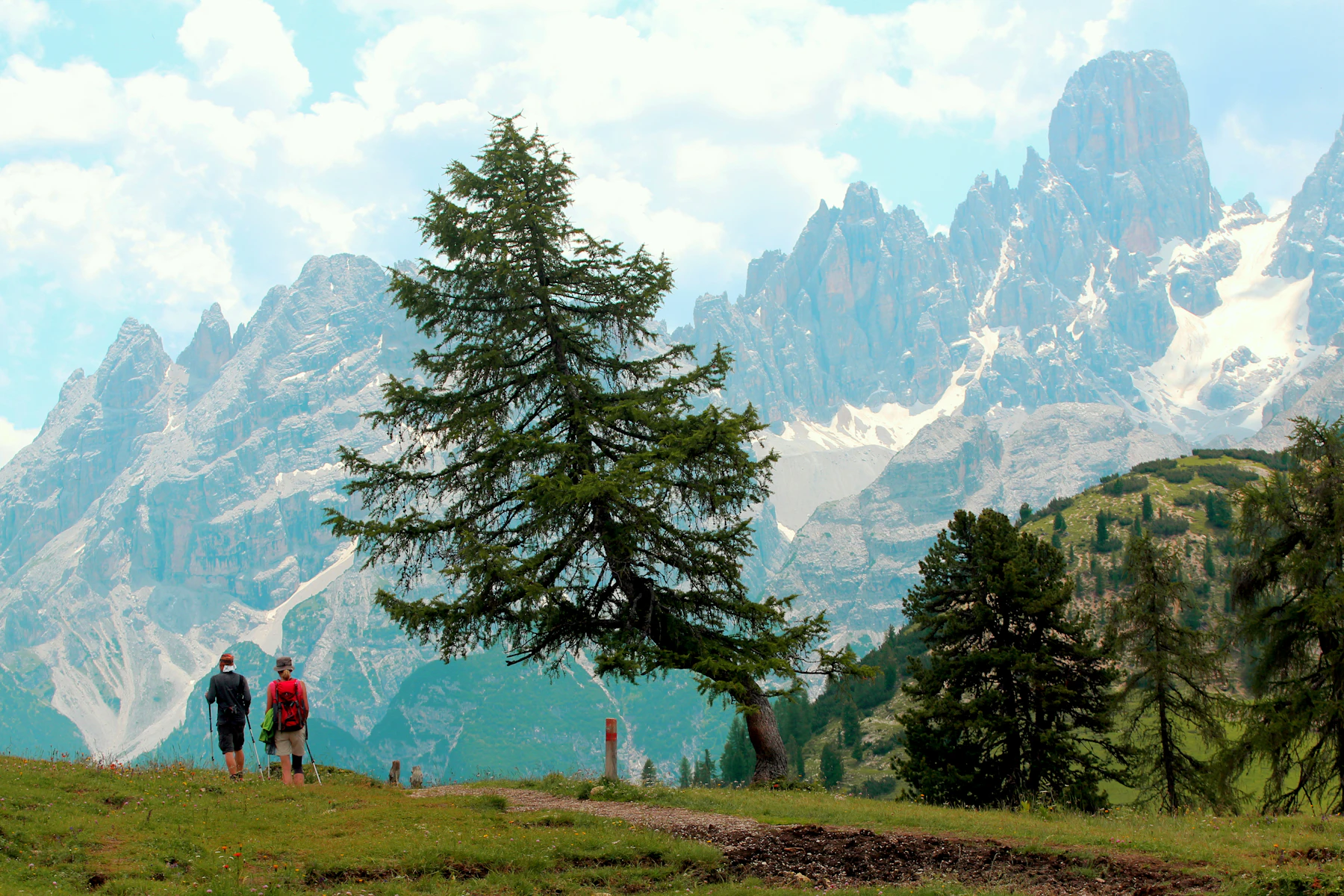 Photograph of Two hikers cross a green Dolomites meadow towards a wall of pale peaks.