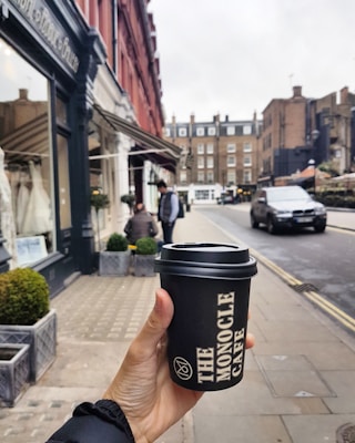 A hand holding a black takeaway coffee cup with the logo 'The Monocle Cafe' printed on it, set against a street scene with red-bricked buildings, shops with awnings, and a moving car on the road. The street is lined with small plants in square pots.