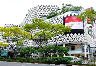 A modern building with a unique, hexagonal-patterned facade featuring green plants integrated into the design. The building has large, prominent advertisements and storefronts including Uniqlo. Several vibrant green trees with yellowish leaves line the street in front of the building. A traffic sign is visible, directing towards Middle Road and Beach Road. Cars are parked or moving by the roadside.