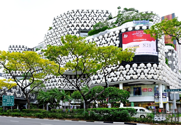 A modern building with a unique, hexagonal-patterned facade featuring green plants integrated into the design. The building has large, prominent advertisements and storefronts including Uniqlo. Several vibrant green trees with yellowish leaves line the street in front of the building. A traffic sign is visible, directing towards Middle Road and Beach Road. Cars are parked or moving by the roadside.