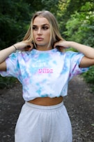 A person stands outdoors wearing a tie-dye shirt with the words 'vibe' printed on it. The background features lush greenery, suggesting a forest or park setting.