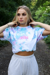 A person stands outdoors wearing a tie-dye shirt with the words 'vibe' printed on it. The background features lush greenery, suggesting a forest or park setting.