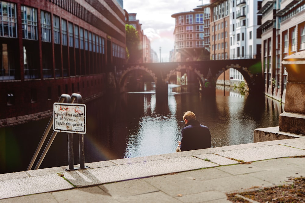 man in black jacket sitting on concrete dock near river during daytime