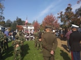 A group of people gathered outdoors at what appears to be a formal event. Several individuals are wearing military uniforms, including camouflage and dress uniforms. American flags are prominently displayed, and the setting is surrounded by autumn trees with colorful leaves. The event seems to be taking place in a residential area with houses visible in the background.
