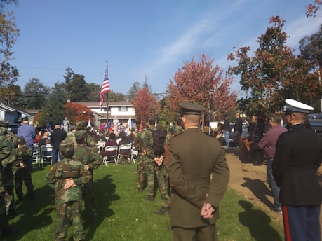 A group of people gathered outdoors at what appears to be a formal event. Several individuals are wearing military uniforms, including camouflage and dress uniforms. American flags are prominently displayed, and the setting is surrounded by autumn trees with colorful leaves. The event seems to be taking place in a residential area with houses visible in the background.