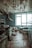 An abandoned kitchen with peeling paint on the ceiling and walls covered in blue tiles. The room features old and rusty kitchen appliances, a broken chair, and a sink. The floor is tiled, and a bright window casts light into the neglected space.