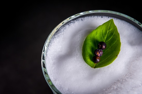 A close-up of a frothy beverage in a clear glass garnished with a vibrant green leaf and three small dark berries.