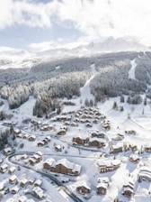 A snow-covered mountain resort featuring numerous wooden chalets nestled among alpine trees. The landscape is dominated by the vast, rolling mountains in the background, offering a picturesque winter scene. Winding roads connect the buildings, with ski tracks visible on the slopes.