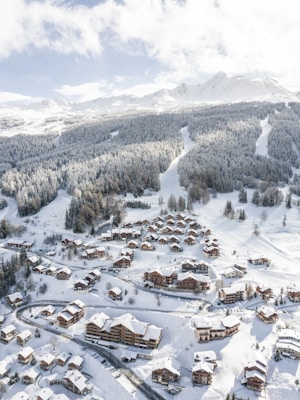 A snow-covered mountain resort featuring numerous wooden chalets nestled among alpine trees. The landscape is dominated by the vast, rolling mountains in the background, offering a picturesque winter scene. Winding roads connect the buildings, with ski tracks visible on the slopes.