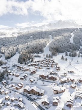 A snow-covered mountain resort featuring numerous wooden chalets nestled among alpine trees. The landscape is dominated by the vast, rolling mountains in the background, offering a picturesque winter scene. Winding roads connect the buildings, with ski tracks visible on the slopes.