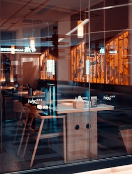 An empty restaurant setting in warm lighting with wooden chairs and tables, seen through a glass window. The interior features hanging pendant lights and decorative wall panels with abstract designs. Reflections are visible on the glass, adding depth to the scene.