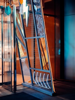 A modern and artistic doorway with geometric shapes and a metallic finish. The door features a prominent lightning bolt design, illuminated sections, and a mix of mirrored and transparent panels. Hanging decorations include teal and white geometric shapes. The interior reflects warm, amber lighting contrasting with the cooler, blue tones of the door.