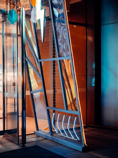 A modern and artistic doorway with geometric shapes and a metallic finish. The door features a prominent lightning bolt design, illuminated sections, and a mix of mirrored and transparent panels. Hanging decorations include teal and white geometric shapes. The interior reflects warm, amber lighting contrasting with the cooler, blue tones of the door.