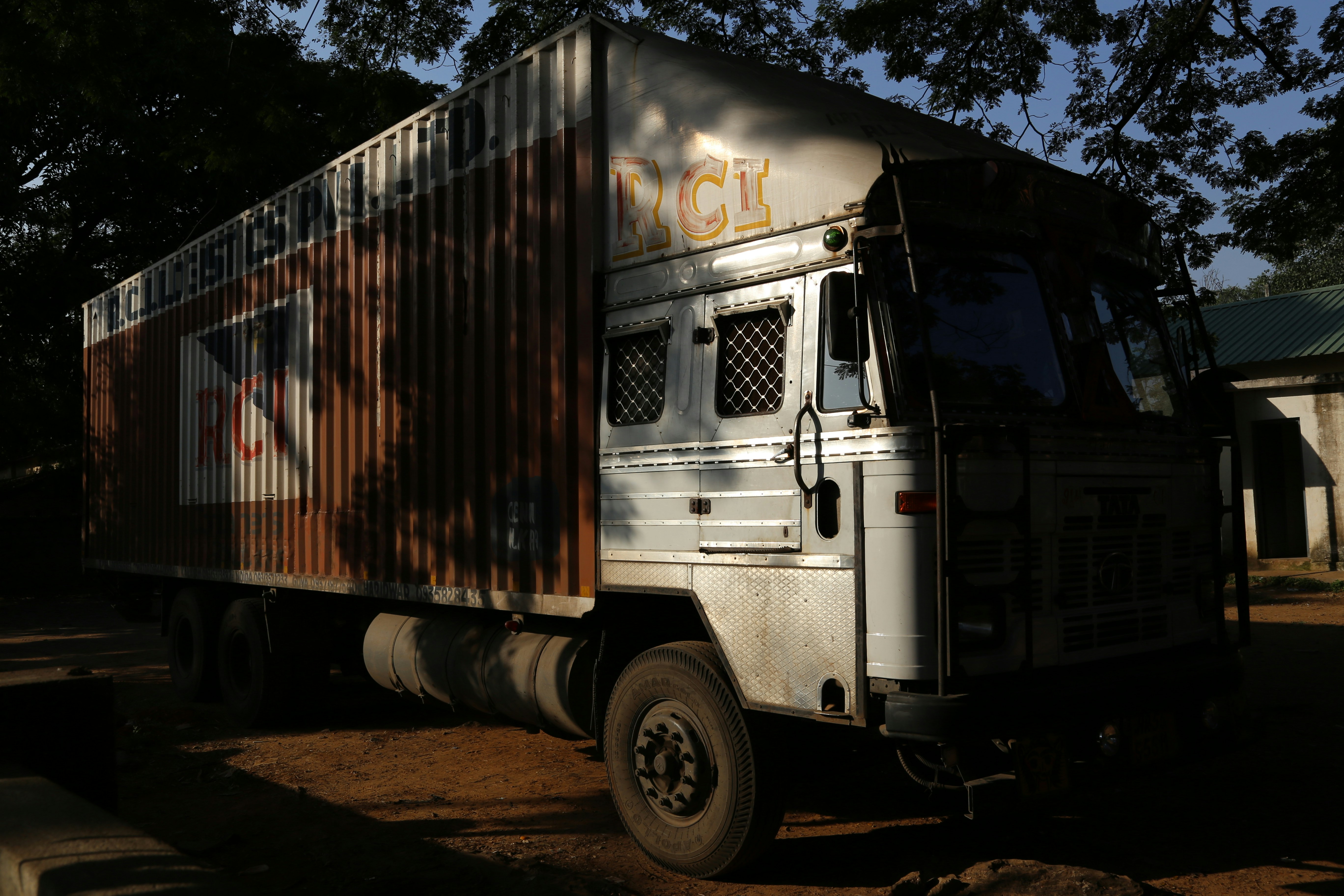 A large truck with a shipping container body parked in a shady area, surrounded by tall trees. The truck's cabin is metallic with a grille on the window, and the container has branding and signage on the side.