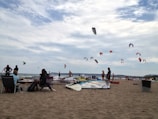 A beach scene with several people enjoying various activities. Many colorful kites are flying in the sky, indicating kite surfing or kitesurfing. People are sitting on beach chairs, and windsurfing boards are visible on the sand. The background shows a vast expanse of calm ocean under a partly cloudy sky.