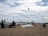 A beach scene with several people enjoying various activities. Many colorful kites are flying in the sky, indicating kite surfing or kitesurfing. People are sitting on beach chairs, and windsurfing boards are visible on the sand. The background shows a vast expanse of calm ocean under a partly cloudy sky.