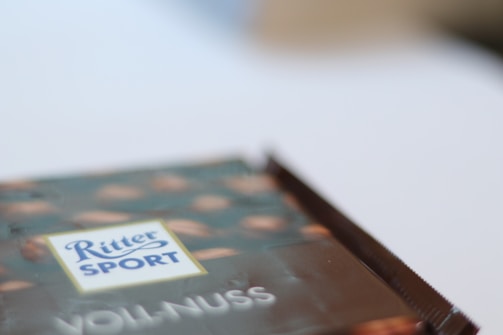 A blurred image focusing on a chocolate packaging with the words 'Ritter Sport' and 'Voll-Nuss' visible. The packaging design includes a pattern of nuts on a dark background.