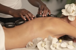 A person is lying face down with smooth black stones placed along their spine. Another person's hands are arranging the stones. Orchid flowers are present, adding a serene and natural touch to the scene. The setting suggests a spa or massage environment focused on relaxation.