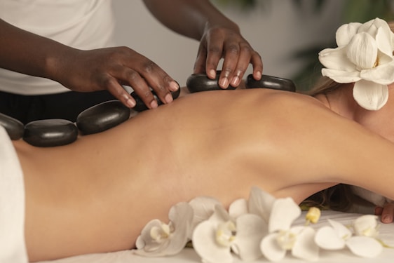 A person is lying face down with smooth black stones placed along their spine. Another person's hands are arranging the stones. Orchid flowers are present, adding a serene and natural touch to the scene. The setting suggests a spa or massage environment focused on relaxation.