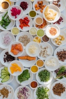 A variety of small plates with colorful, diverse food items including vegetables, sauces, cheeses, and meats. Each plate contains a different ingredient, such as sliced cucumbers, beet hummus, sun-dried tomatoes, herbs, and seeds. The arrangement is vibrant and visually appealing.