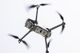 A drone captured in flight against a light sky background. The focus is on the body and propellers, with rotors in motion creating a blur effect. The drone appears sleek and modern, featuring sensors and a camera on the front.