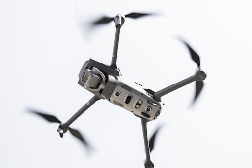 A drone captured in flight against a light sky background. The focus is on the body and propellers, with rotors in motion creating a blur effect. The drone appears sleek and modern, featuring sensors and a camera on the front.