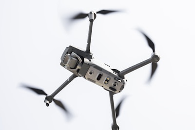 A drone captured in flight against a light sky background. The focus is on the body and propellers, with rotors in motion creating a blur effect. The drone appears sleek and modern, featuring sensors and a camera on the front.