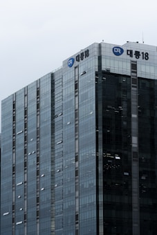 A tall, modern glass skyscraper with reflective windows and a logo near the top of the building. The architecture features a grid-like pattern of windows with some open for ventilation.