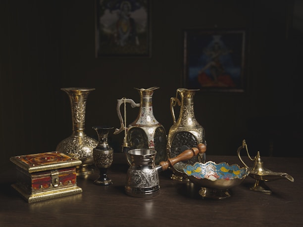 A collection of vintage brass and silverware items arranged on a table. The set includes intricately designed vases, pitchers, a traditional lamp, a small ornate box, and a decorative bowl. The background is dark, highlighting the shine and textures of the metal objects.
