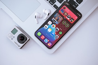 A smartphone with a colorful home screen displaying various app icons is lying on a closed laptop keyboard. Next to it are a pair of wireless earphones and a compact action camera. The composition highlights modern digital technology gadgets arranged on a flat surface.