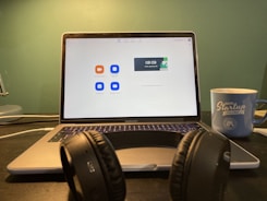 A laptop displaying a video conferencing application screen is placed on a desk. In front of the laptop, there is a pair of black headphones resting on the desk. To the right, a blue mug with 'Digital Startup Challenge' printed on it is positioned. The background wall is green, creating a subtle contrast with the objects in the foreground.